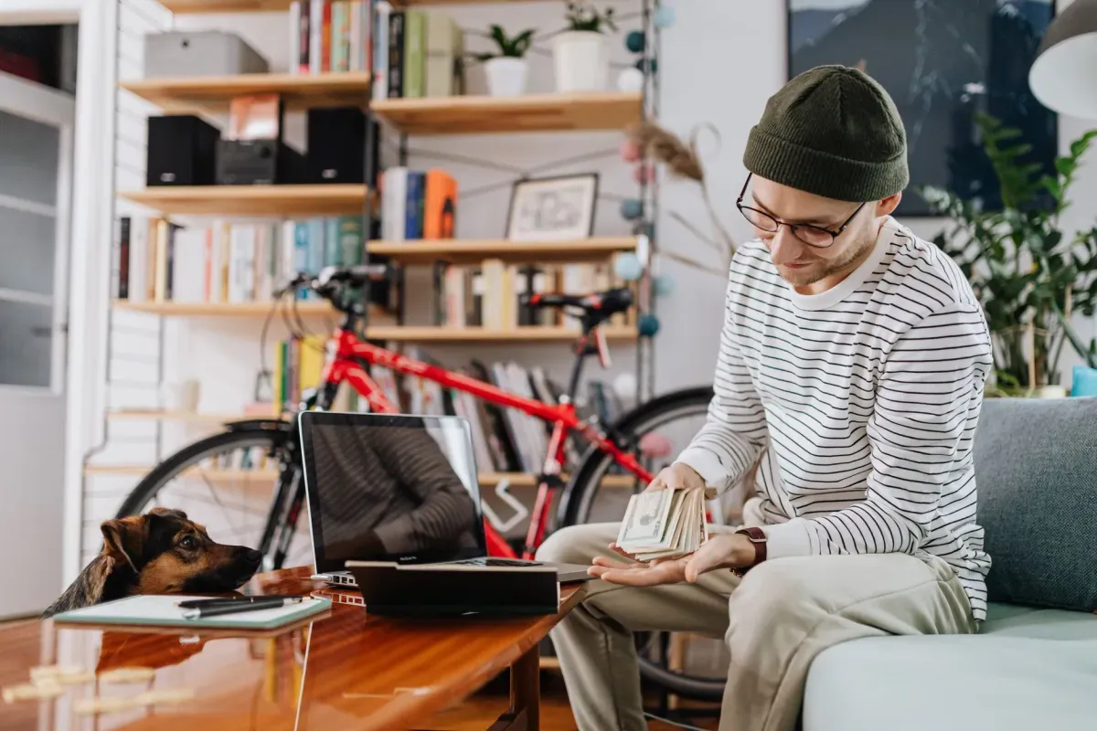 Homem sentado no sofá com o notebook ao lado, contando o dinheiro para organizar as finanças de casa.