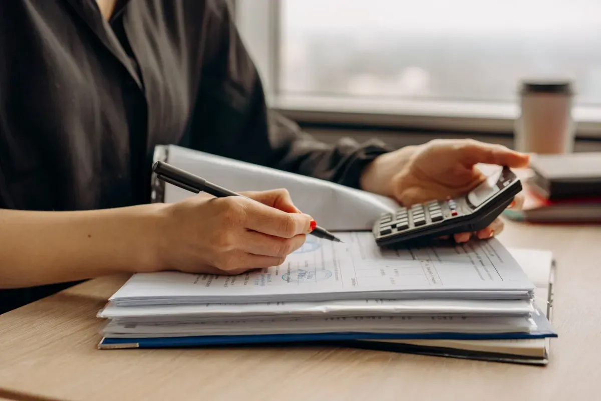 Mãos fazendo contas na calculadora e anotando com caneta sobre uma pilha de documentos