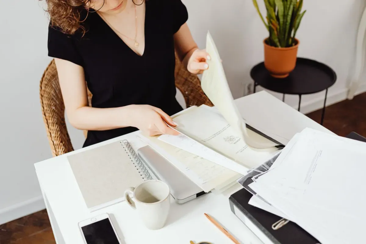Mulher separando documentos e pastas na mesa de trabalho, com caderno, celular e caneca