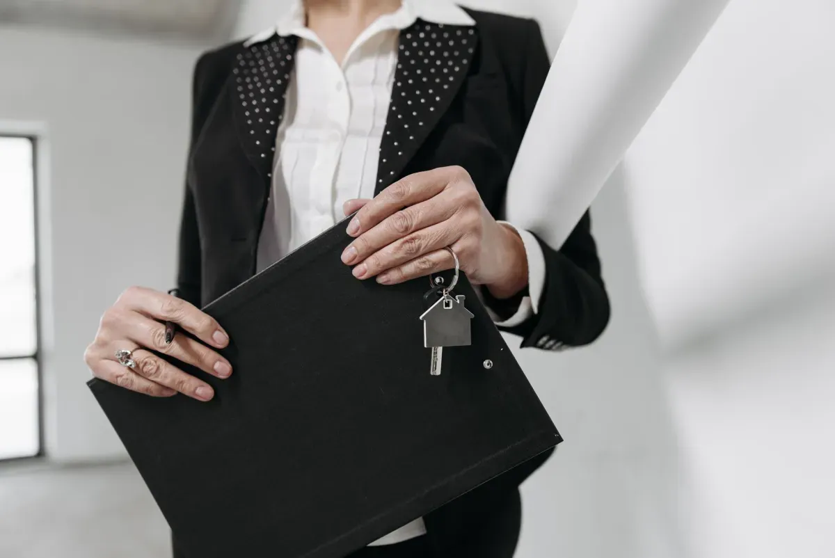 Mulher de blazer segurando pasta preta e chaveiro em formato de casa