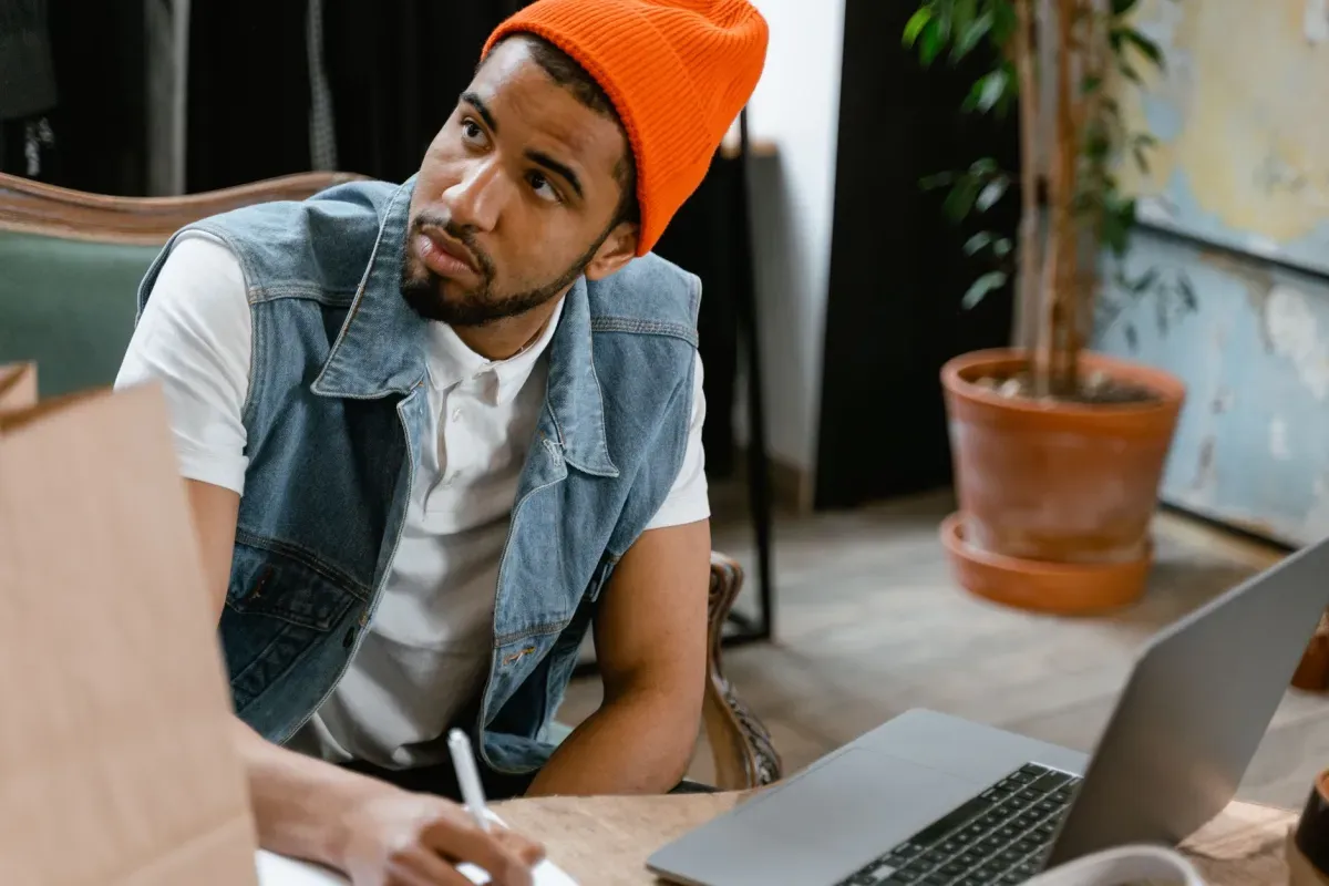 Homem de gorro laranja pensativo enquanto trabalha no notebook