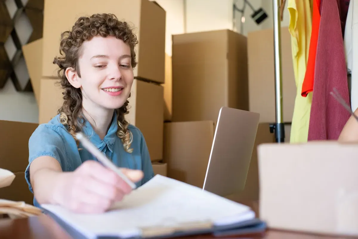 Jovem empreendedora anotando pedidos entre caixas de envio e araras de roupa