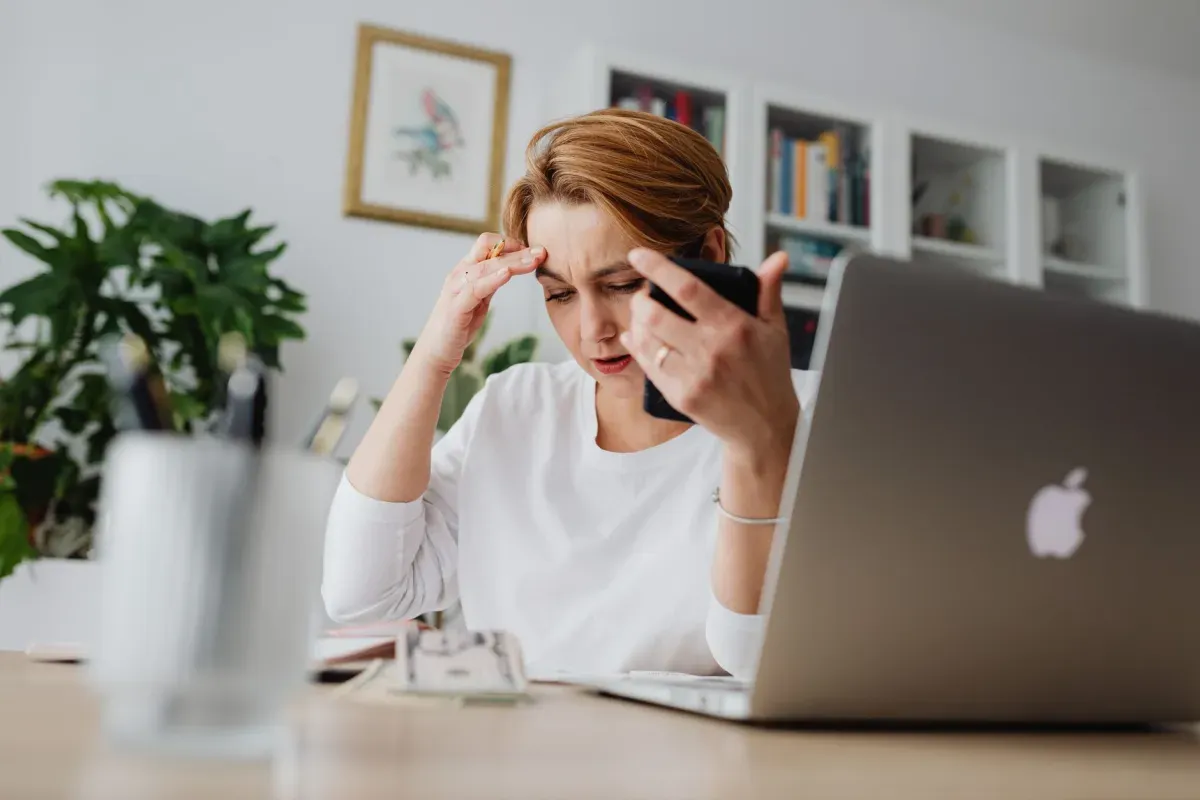 Mulher preocupada ao celular diante do notebook e de papéis na mesa