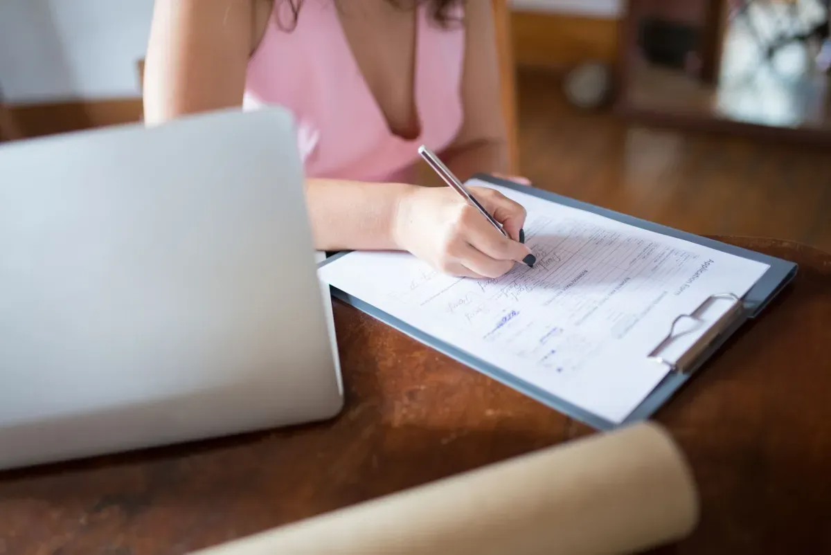 Mulher escrevendo em um documento na prancheta, com notebook ao lado